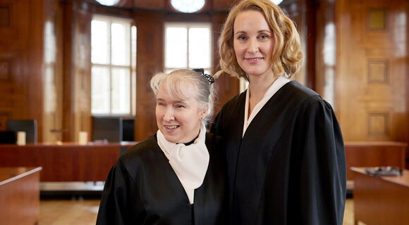 Pamela Pabst und Christina Athenstädt, beide in schwarzer Robe, stehen in einem  Gerichtssaal, Pamela Pabst trägt ihr helles Haar hochgesteckt.