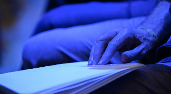 Praktisch und kreativ: eine Hand fährt beim blauen Licht eines Konzerts über ein Liederbuch in Braille-Schrift.