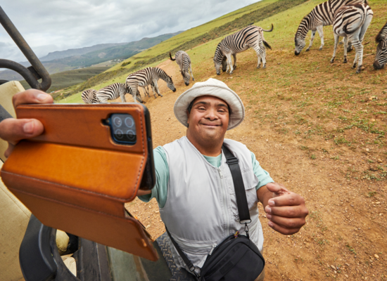 Ein Mann mit gebräunter Haut und einem weißen Hut steht vor ein paar Zebras. Er lächelt. In seiner Hand hält ein Smartphone vor sich als würde er ein Selfie machen. Neben ihm steht ein Geländewagen.