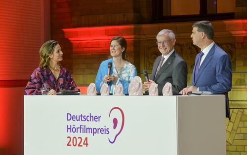 Auf der Bühne hinter einem weißen Pult mit dem Logo des Deutschen Hörfilmpreises stehen folgende Personen von links nach rechts: Nadine Heidenreich, Dr. Ulrike Köhler, Bundespräsident a. D. Prof. Dr. Horst Köhler und Dr. Thomas Krämer