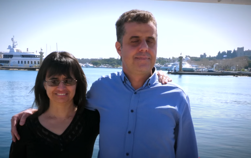Links steht eine Frau mit mittellangen dunklen Haaren und dunkler Sonnenbrille. Neben ihr ein Mann mit kurzen dunklen Haaren und geschlossenen Augen. Er legt den Arm um sie. Yachten, Wasser und blauer Himmel im Hintergrund.