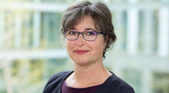 Porträt von Julia Gebrande: Eine Frau mit kurzem braunem Haar und Brille blickt in die Kamera. Sie trägt ein dunkles Jackett über einem gemusterten Oberteil. Der Hintergrund ist hell und unscharf.