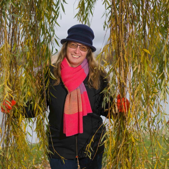 Kathleen Starke, umrahmt von den Ästen und Blättern einer Trauerweide. Sie hat langes helles Haar und trägt eine getönte Brille. Mütze und Jacke sind schwarz, ihr großer Schal ist pink-lila-gestreift.