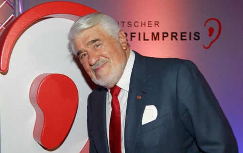 Mario Adorf, ein älterer grauhaariger Mann, steht lächelnd vor einem Plakat mit der Aufschrift "Deutscher Hörfilmpreis". Er trägt Anzug und Krawatte.