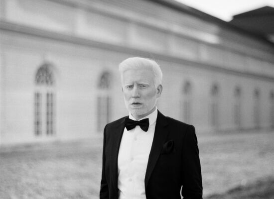 Marschall Alam steht in einem schwarzem Anzug und einem weißen Hemd mit schwarzer Fliege vor einem historischen Gebäude.  Er hat weißes Haar und einen Dreitagebart.