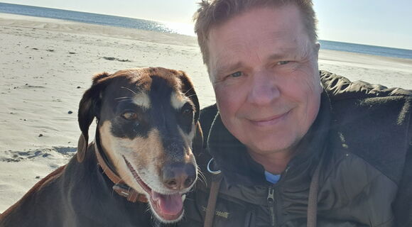 Ein Mann mit kurzen, hellen Haaren und einer dunklen Jacke macht ein Selfie mit einem braun-schwarzen Hund am Sandstrand. Beide schauen freundlich in die Kamera. Im Hintergrund sind Meer, blauer Himmel und Sonnenlicht zu sehen.