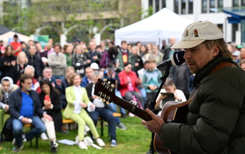 Ein Blick von der Außentribüne auf dem Berliner Platz ins Publikum der Eröffnungsfeier: rechts im Bildvordergrund steht scharf zu sehen Philipp Poisel mit seiner Gitarre, den leicht verschwommenen Besucherinnen und Besuchern zugewandt.