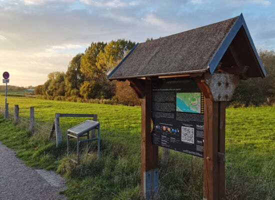 Am Wegesrand steht eine Infotafel zum Planeten Erde, daneben ist die entsprechende Station des Planetenlehrpfades in Wedel. Der Weg führt weiter in die Ferne, umgeben von Wiesen.