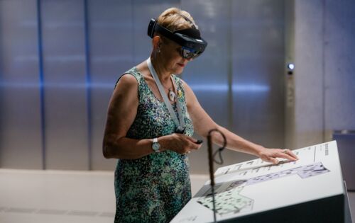 Eine blinde Frau mit kurzen Haaren und Langstock bei der HoloAudioTour im Deutschen Museum Nürnberg. Sie trägt eine AR-Brille, in der einen Hand hält sie eine Fernbedienung. Mit der anderen Hand ertastet sie eine Erklärtafel.