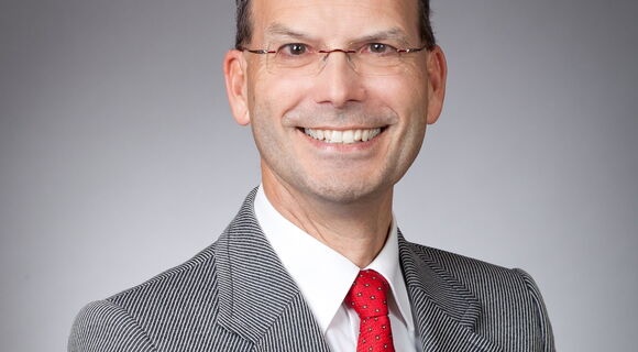 Stefan Insam lächelt in die Kamera. Er hat kurzes Haar, und trägt eine kleine randlose Brille. Unter einem Sakko trägt er ein weißes Hemd und eine Krawatte.