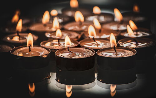 Viele Teelichter stehen auf einer spiegelnden Oberfläche. Das Licht der Flammen der vorderen drei Lichter spiegelt sich darin.