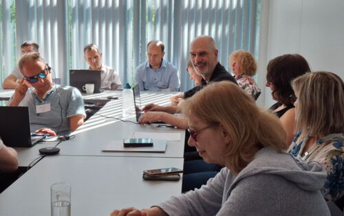 Beim Workshop „Überregionale Kooperation“: Teilnehmerinnen und Teilnehmer sitzen an Tischen. Vor manchen liegt ein Smartphone, andere haben ihr Laptop aufgeklappt. Auch Workshop-Leiter Reiner Delgado bedient ein Laptop, während er lächelnd Richtung Kamera