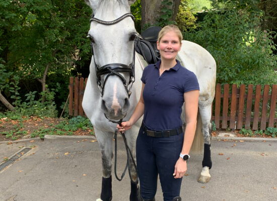 Helen Vogt steht mit ihrer Schimmelstute Janita auf einem Reitplatz, der von Büschen und Bäumen umgeben ist. Sie hat langes blondes, zu einem Pferdeschwanz gebundenes Haar und trägt ein kurzärmeliges Polohemd. Janita trägt einen Sattel und Zaumzeug.