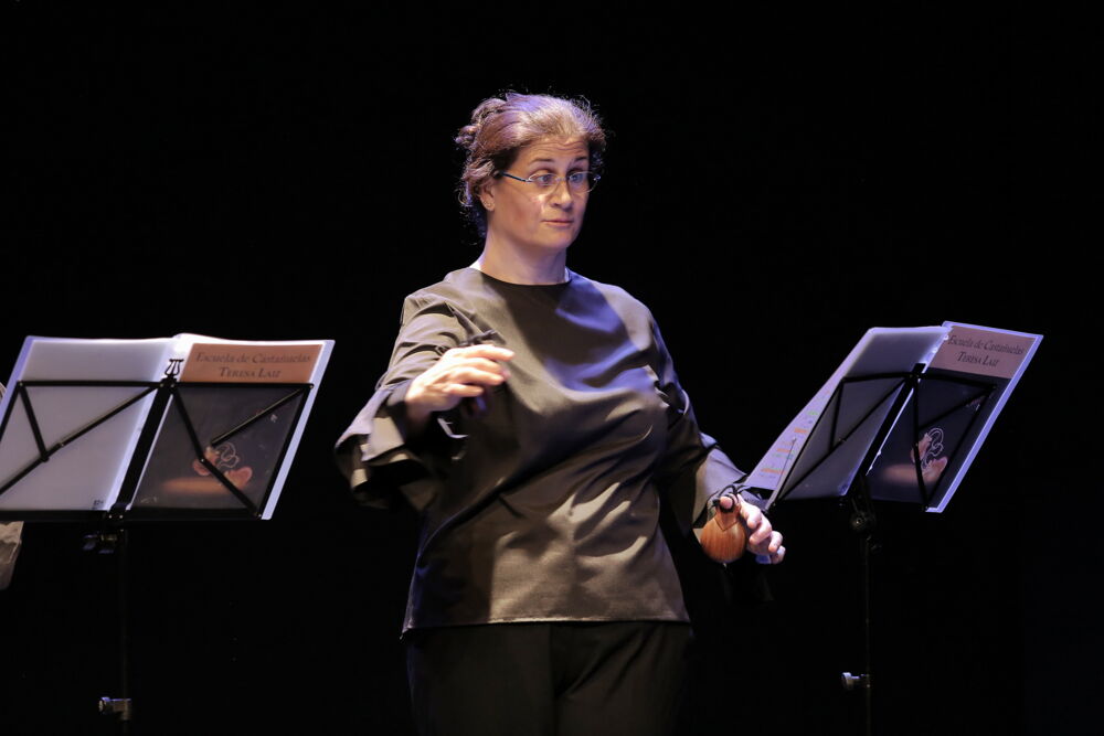 Bárbara Martín Muñoz steht auf einer Bühne, mit beiden Händen spielt sie je eine Kastagnette. Sie blickt auf Noten auf einem Notenständer. Ihr Haar hat sie hochgebunden, sie trägt eine Brille.