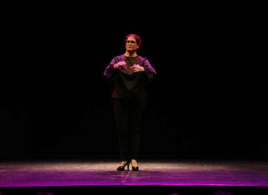 Ganzkörperbild von Bárbara Martín Muñoz auf einer dunklen Bühne. Mit beiden Händen spielt sie Kastagnetten. Sie trägt eine Brille und hat dunkle Kleidung und hohe Schuhe an.