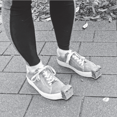 Ein Paar Beine in schwarzen Leggings und mit den besonderen Schuhen: An deren Spitze ist jeweils das InnoMake-Gerät befestigt, ein kleiner Kasten aus Metall.