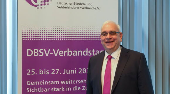 DBSV-Präsident Hans-Werner Lange steht lächelnd vor einem Plakat, das auf den Verbandstag 2026 hinweist. Er hat kurzes graues Haar, trägt eine getönte Brille, Anzug und Krawatte.