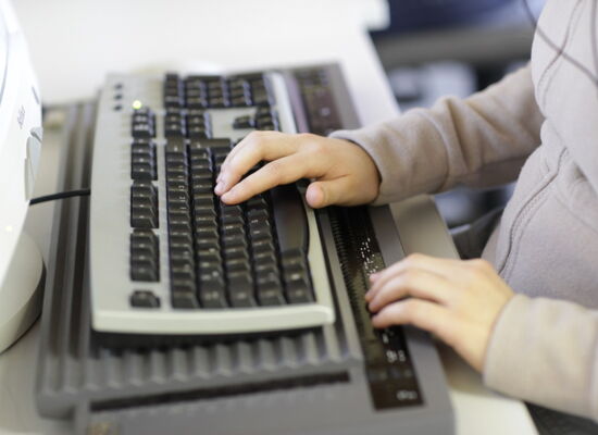 Eine Person arbeitet an einem Computer mit einer Braillezeile und Tastatur. Die linke Hand bedient die Tastatur, während die rechte Hand die taktilen Informationen auf der Braillezeile ertastet