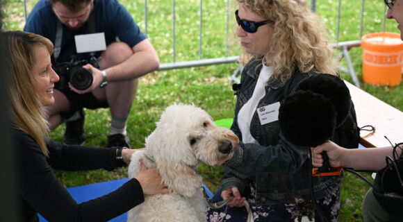 Zwei junge Frauen sitzen sich gegenüber, zwischen ihnen ein heller Goldendoodle. Ein Mann filmt die Szene.