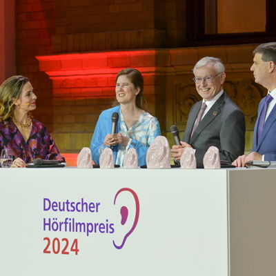 Moderatorin Nadine Heidenreich, Dr. Ulrike Köhler, Bundespräsident a. D. Prof. Dr. Horst Köhler und Dr. Thomas Krämer stehen in festlicher Kleidung hinter einem Pult, auf dem "Deutscher Hörfilmpreis 2024" steht.