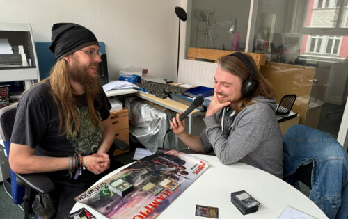Felix Högl und Christian Balbach sitzen gemeinsam an einem Tisch, auf dem Spielkarten von Magic: The Gathering sowie eine Spielunterlage liegen. Beide sind ins Gespräch vertieft und lachen.