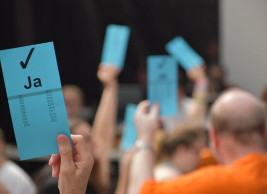 Bei einer Abstimmung hält eine Hand einen blauen Zettel mit dem Wort „Ja“ und einem Haken nach oben. Im Hintergrund sind mehrere Personen zu sehen, die das gleiche tun.
