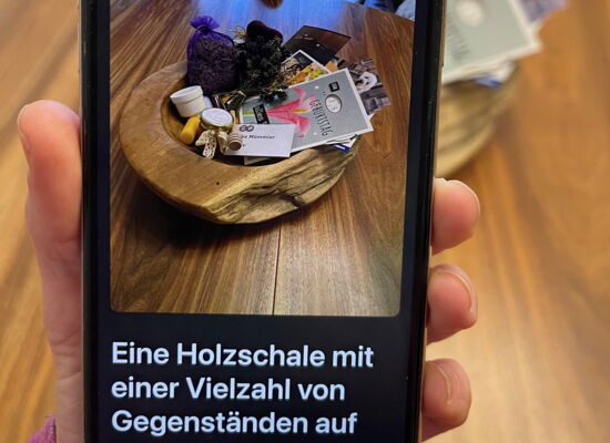 Auf einem Smartphonedisplay ist durch den Kamerasucher eine Deko-Schale zu sehen, in der ein Lavendelsäckchen, Karten und ein Namenschild liegen. Darunter steht: "Eine Holzschale mit einer Vielzahl von Gegenständen auf..."