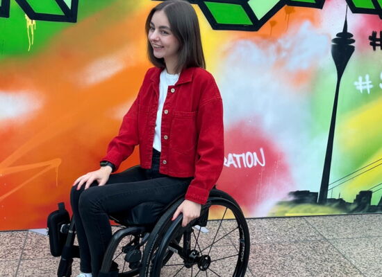 Katharina Hesener sitzt in einem Rollstuhl. Sie ist schlank und hat mittellanges dunkles Haar. Sie trägt eine modische kurze rote Jacke, dunkle Jeans und weiße Sneakers.
