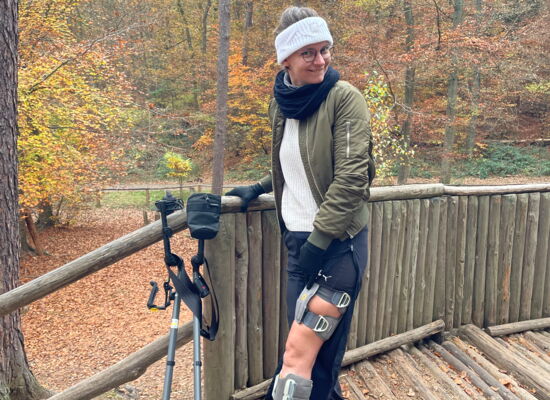 Lea Menninger steht auf einem Bohlenweg an einem Holzgeländer. Am Bein trägt sie ein FES-System, mit dessen Hilfe Nerven angeregt werden.