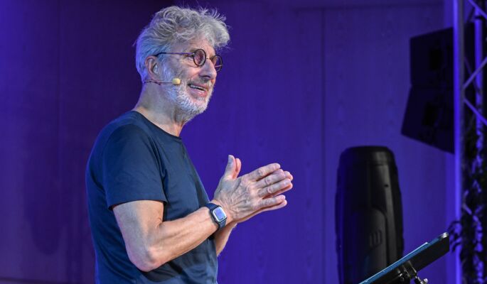 Der Oberkörper von Rufus Beck im Profil vor einem Stehpult, im Hintergrund eine blau angeleuchtete Holzwand und Lautsprecher. Er hat kurze graue Haare, trägt ein dunkles T-Shirt, eine Brille und eine Armbanduhr. An seiner Wange ist ein Mikrofon befestigt.