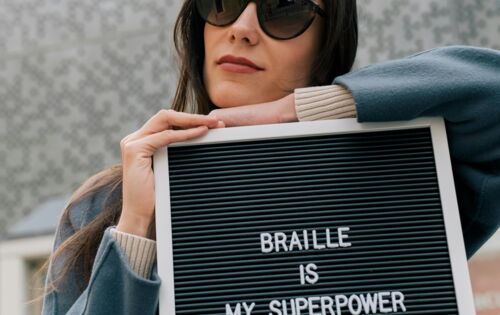 Eine Frau mit braunen langen Haaren lehnt sich auf ein Schild auf dem „BRAILLE IS MY SUPERPOWER“ steht. Sie hat einen blauen Mantel an und trägt eine dunkle Brille.