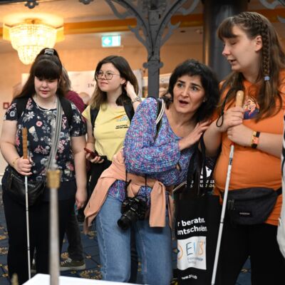 Vier Frauen stehen interessiert vor einem Stand. Eine von ihnen, eine Frau mit dunklen Haaren und einer Kamera um ihren Hals, hat erstaunt den Mund geöffnet. Sie lehnt sich zu einer jungen Frau neben ihr. Diese trägt ein oranges T-Shirt, hat eine Flechtfr