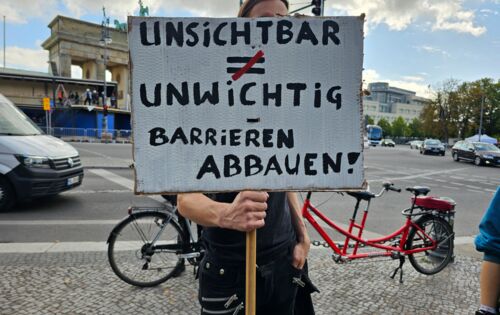 Ein Mann hält ein Schild in die Kamera: „UNSICHTBAR ≠ UNWICHTIG – BARRIEREN ABBAUEN“. Er steht vor dem Brandenburger Tor. Im Hintergrund ist ein rotes Tandem zu sehen.