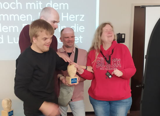 Bei der Preisverleihung nehmen Lucas Brinse, Reiner Delgado und Annette Sander den Preis entgegen. Sie halten die Skulptur gemeinsam in den Händen: Das Herz aus hellem Holz, auf dem ein Auge und ein Ohr zu sehen sind, steht auf einem Sockel.