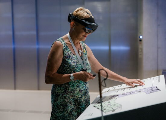 Eine blinde Frau mit kurzen Haaren und Langstock bei der HoloAudioTour im Deutschen Museum Nürnberg. Sie trägt eine AR-Brille, in der einen Hand hält sie eine Fernbedienung. Mit der anderen Hand ertastet sie eine Erklärtafel.