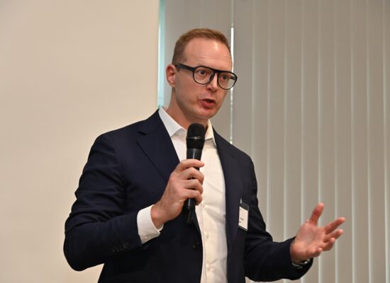 Professor Robert Finger spricht in ein Mikro, das er in der Hand hält. Er hat kurzes Haar, trägt eine große Brille mit dunklem Rahmen, ein dunkles Sakko und ein weißes Hemd.