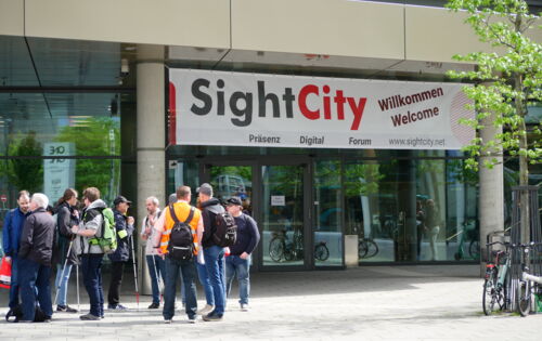 : Vor dem Eingang der Sightcity steht eine Gruppe von Menschen. Viele von Ihnen haben einen Langstock dabei. Sie tragen Rucksäcke. Über der Tür hängt ein Willkommens-Banner.
