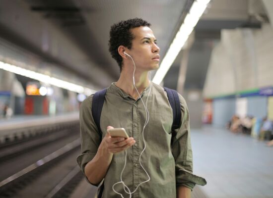 Ein Mann mit dunklen, krausen Haaren steht mit einem Handy in der Hand und Kopfhörern in den Ohren auf einem Bahnsteig. Er blickt mit gerunzelter Stirn irritiert zur Seite. Er trägt ein grünes lockeres Hemd.