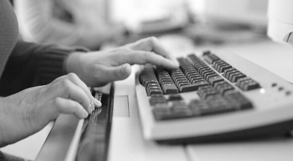 Frauenhände an einer Computertastatur mit Braillezeile. Im Hintergrund ist ein weiterer Arbeitsplatz zu sehen.