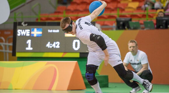 Ein Goalballspieler beim Spiel. Er trägt eine Dunkelbrille und ein weißes Trikot. In seiner linken Hand hält er den Ball bereit zum Wurf.