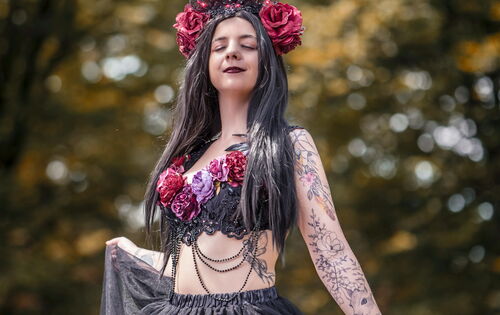 Nadine Rokstein ist eine junge Frau mit langen, schwarzen Haaren. Sie trägt ein schwarzes, mit Rosen verziertes Oberteil und einen langen, schwarzen Tüllrock. Sie hat eine Krone mit roten Rosen auf dem Kopf und trägt ebenfalls florale Armtattoos.  Der Hin
