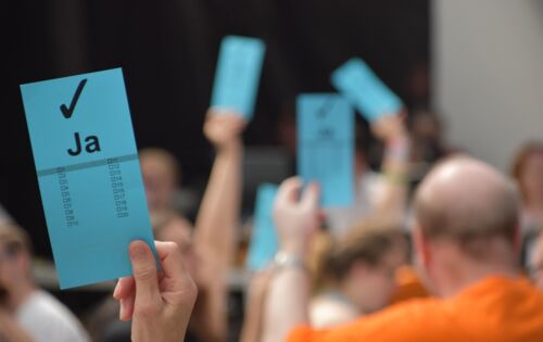 Bei einer Abstimmung hält eine Hand einen blauen Zettel mit dem Wort „Ja“ und einem Haken nach oben. Im Hintergrund sind mehrere Personen zu sehen, die das gleiche tun.