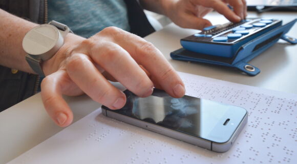 Zwei Hände sind zu sehen: Die linke bedient eine Braillezeile, die rechte ein Smartphone. Das Smartphone liegt auf einer mit Brailleschrift bedruckten Seite.