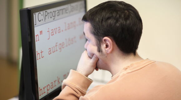 Ein Mann mit kurzen dunklen Haaren blickt, die linke Hand ans Kinn gelegt, nachdenklich auf einen Bildschirm mit stark vergrößerter Schrift.