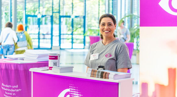 Bei der Jubiläumsfeier in Hessen: Eine Frau steht lächelnd an einem Infostand in einem Gebäude mit Glasfront. Das Rasterauge des DBSV prangt hinter ihr, am Tisch des Standes und auf ihrem T-Shirt.