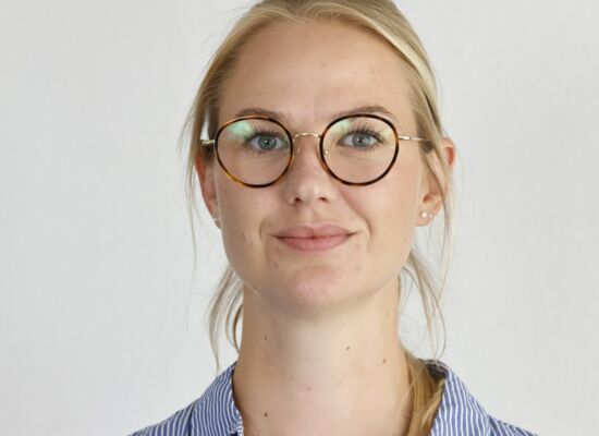 Melissa Glomb hat ihr blondes, glattes Haar zusammengebunden und trägt eine Brille mit runden Brillengläsern und eine helle Hemdbluse.
