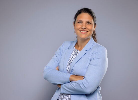 Sarah Karge, eine Frau mit brauen, langen Haaren, steht mit verschränkten Armen vor einem neutralen Hintergrund. Sie trägt einen hellblauen Blazer über einer gemusterten Bluse. Sie lächelt offen in die Kamera.