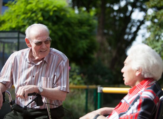 Ein alter Mann und eine alte Frau im Gespräch: Er trägt ein gestreiftes Hemd und stützt sich auf seinen Rollator. Sie sitzt rechts von ihm in einer rot-schwarz-karierten Bluse. Sie sitzen draußen, es ist sonnig.