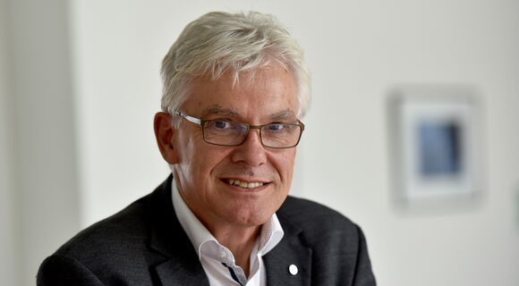 Ein älterer Mann mit weißem Haar, Brille und hellem Teint trägt ein dunkles Sakko über einem weißen Hemd. Er lächelt freundlich und blickt direkt in die Kamera. Der Hintergrund ist neutral und unscharf, mit einem gerahmten Bild an der Wand.