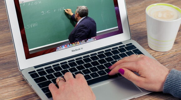 Eine Person tippt auf einem MacBook, auf dessen Bildschirm ein Video mit einem Lehrer läuft, der an eine grüne Tafel schreibt. Die Person trägt graue Kleidung, hat lila lackierte Nägel und trägt Ringe. Daneben stehen eine Tasse Kaffee und ein Smartpho